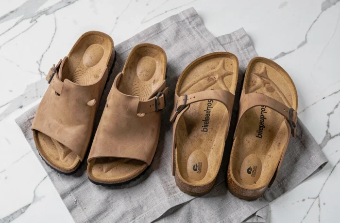 Comparatif visuel entre une chaussure Scholl et une sandale Birkenstock Boston posées côte à côte sur marbre blanc, choix de chaussures de confort